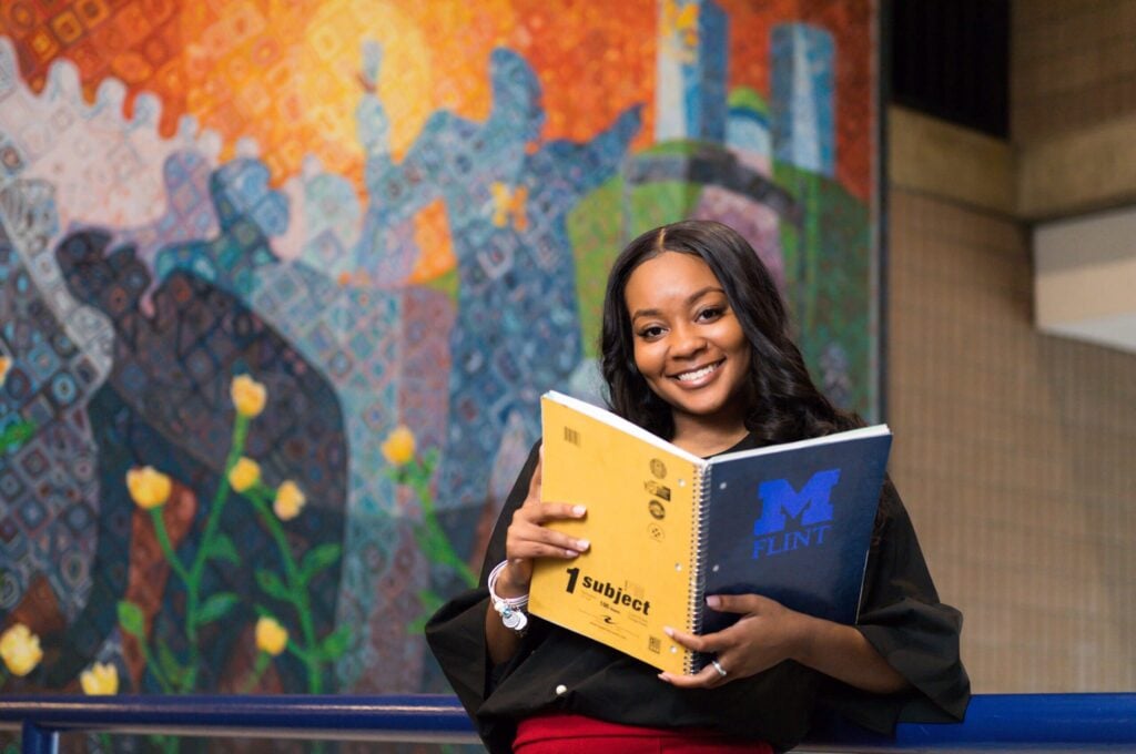 African American female with a UM-Flint notebook in front of a mural