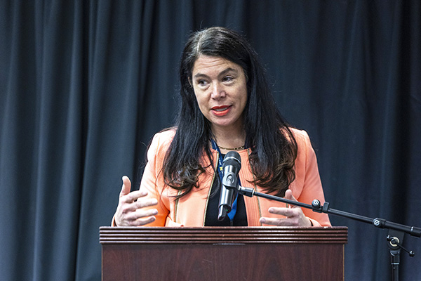 a woman with dark hair and bright jacket at a podium speaking