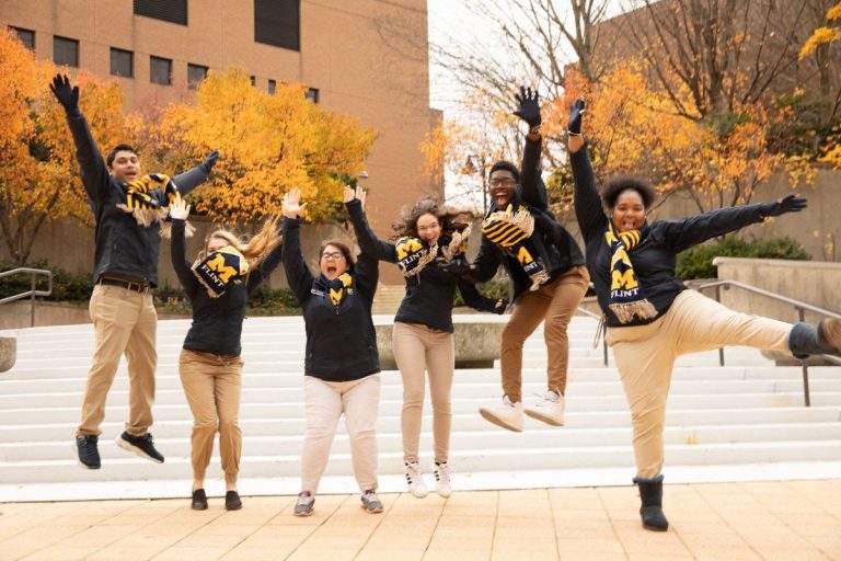 Three college students with University of Michigan-Flint scarves outside jumping into the air