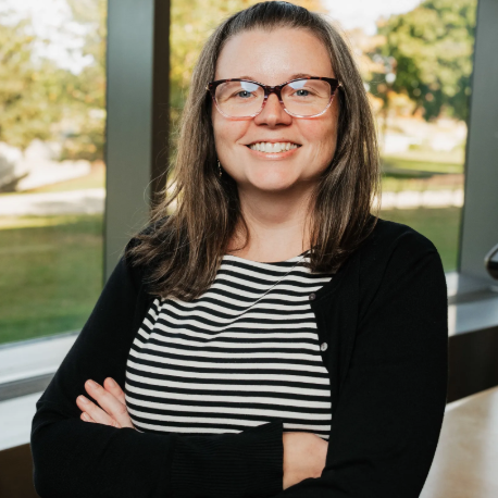 Meagan Brown with glasses smiles while standing with arms crossed in front of large windows overlooking a green landscape.