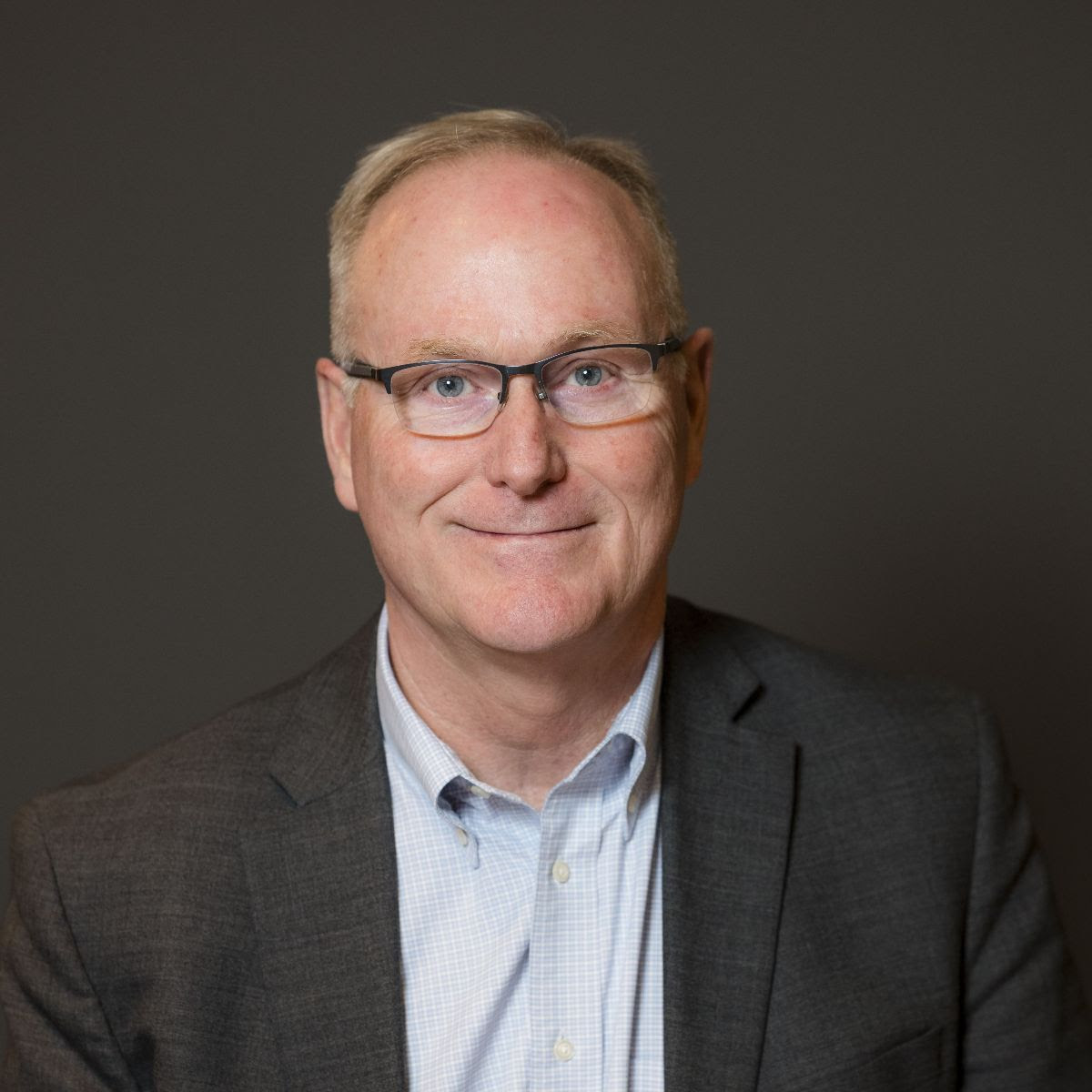 Ken Sylvester wearing glasses and a blazer smiles against a neutral background.