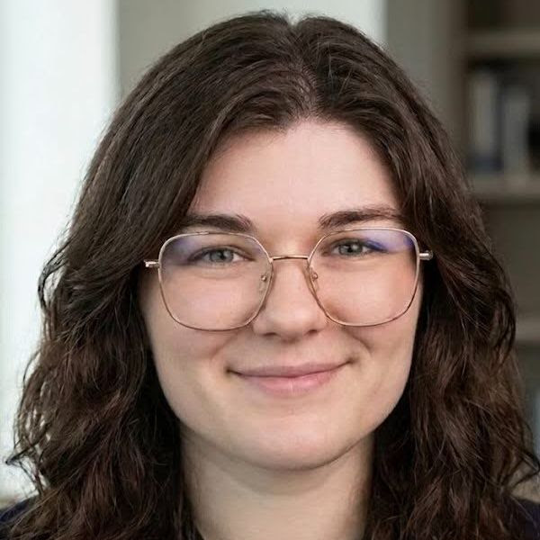 Dakota Allison with glasses smiles at the camera, with a blurred background of shelves.