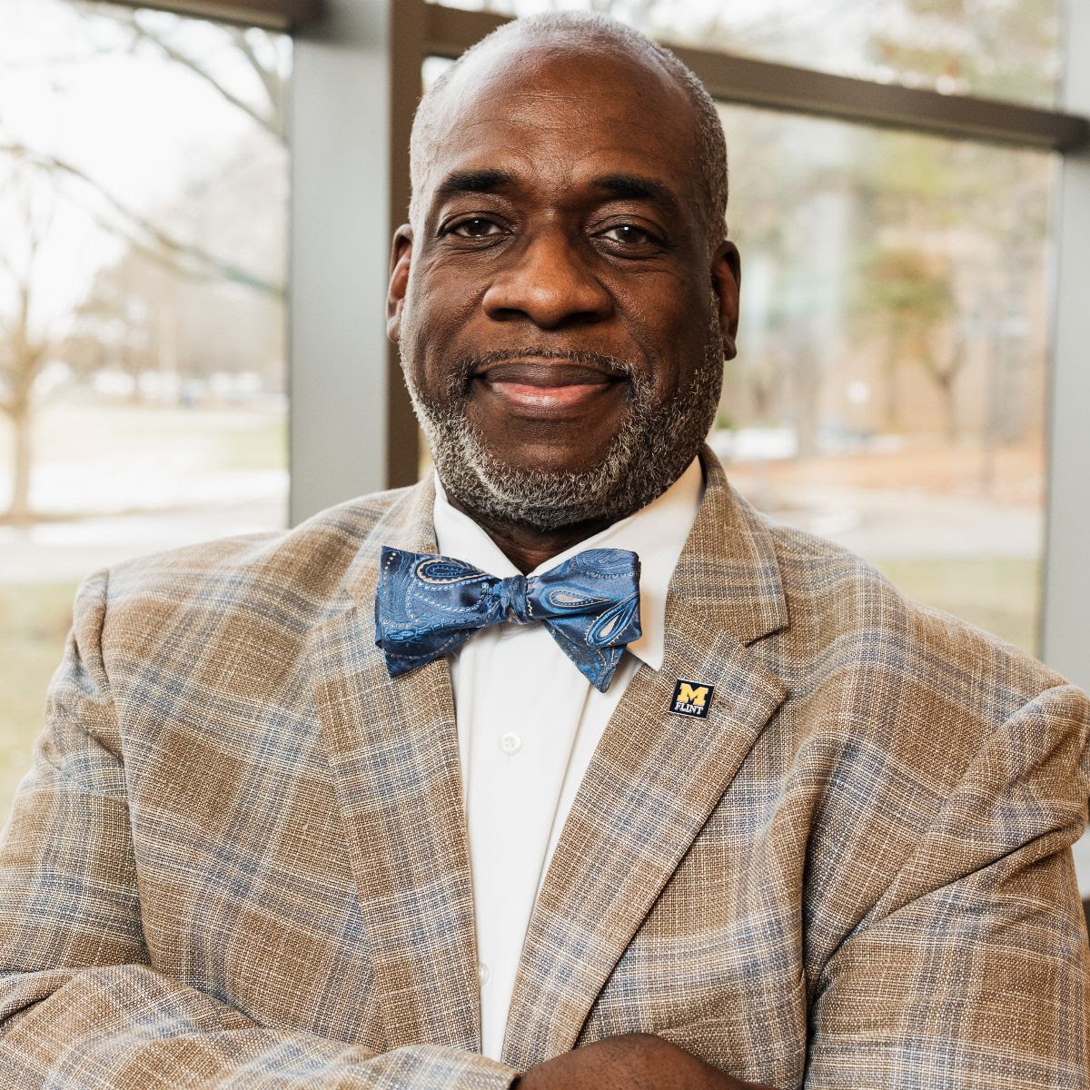 John M. Ambrose in a plaid blazer and bow tie smiles while standing with arms crossed in front of a window.