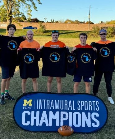 Five people stand together holding black t-shirts, smiling in front of a sign that reads "MICHIGAN FLINT INTRAMURAL SPORTS CHAMPIONS."