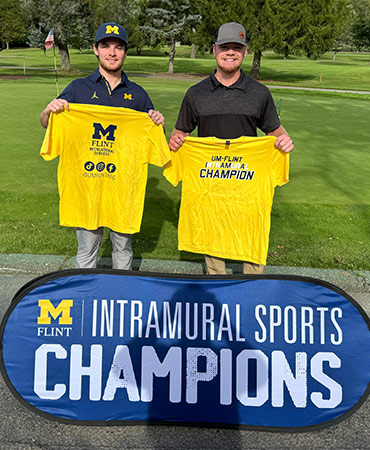 Two people hold yellow t-shirts that say "CHAMPION" while standing in front of a banner that reads "INTRAMURAL SPORTS CHAMPIONS."