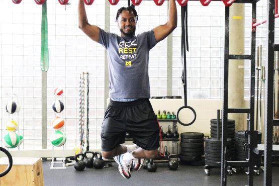 A person hangs from gym rings, demonstrating strength, in a fitness studio with various exercise equipment in the background.