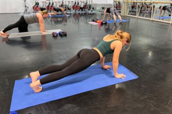 A person performs a plank exercise on a blue mat in a gym, with others exercising in the background.