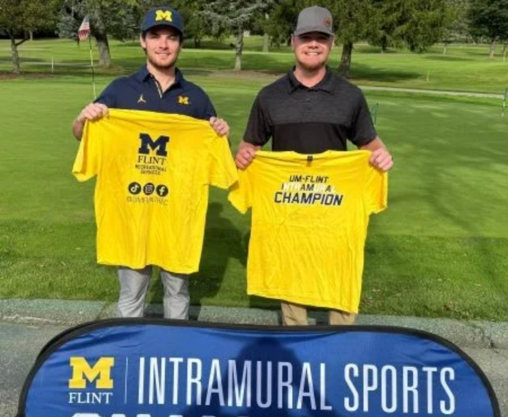Two people stand on a golf course holding yellow t-shirts; one reads "UM-FLINT INTRAMURAL SPORTS" and the other "I AM A CHAMPION."