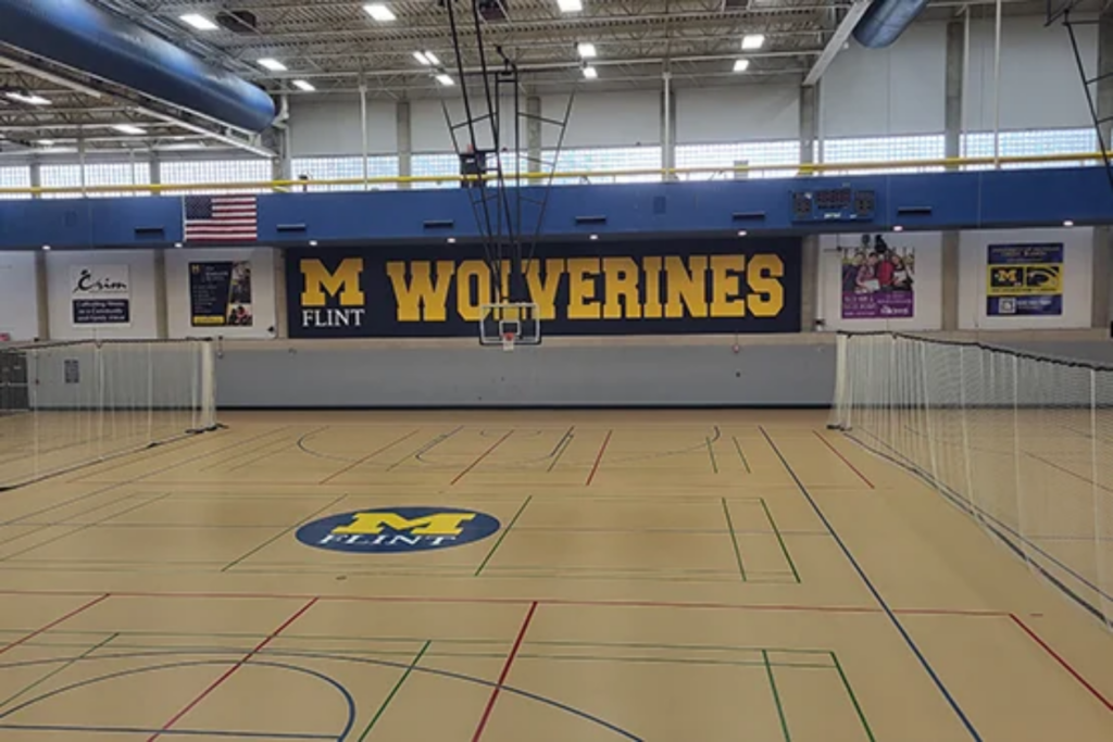A gymnasium with a large "M WOLVERINES" sign on the wall, basketball hoops, and a court marked with colorful lines.