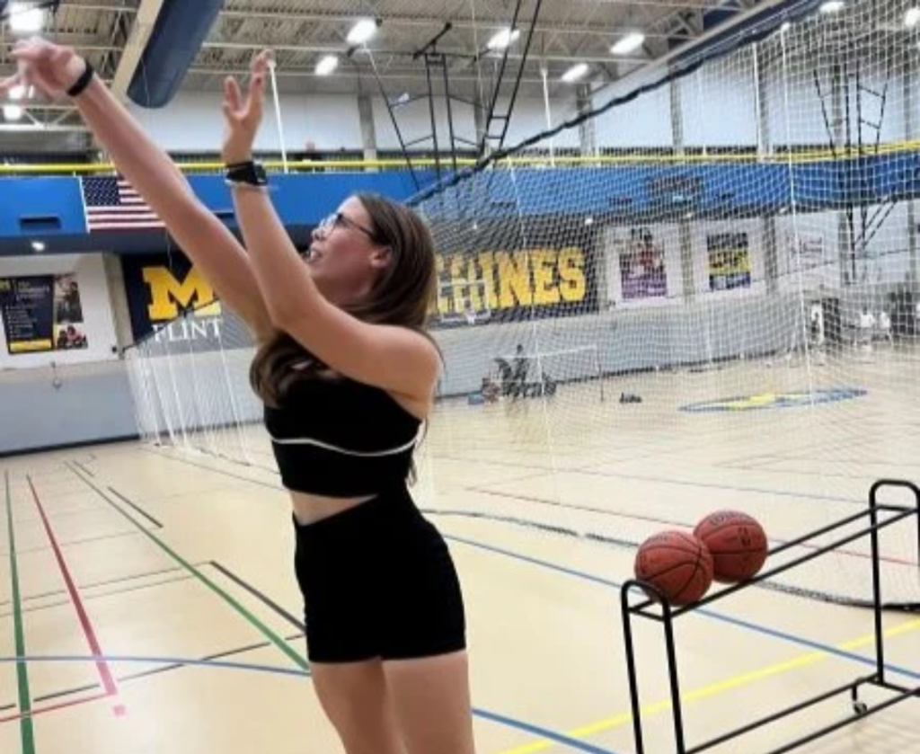 A person is shooting a basketball in a gym with two basketballs on a rack nearby.