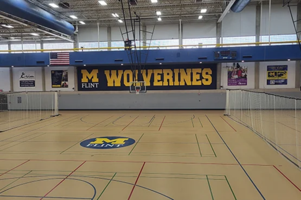 An indoor gymnasium with a basketball hoop, volleyball nets, and a wall displaying "M FLINT WOLVERINES" and an American flag.
