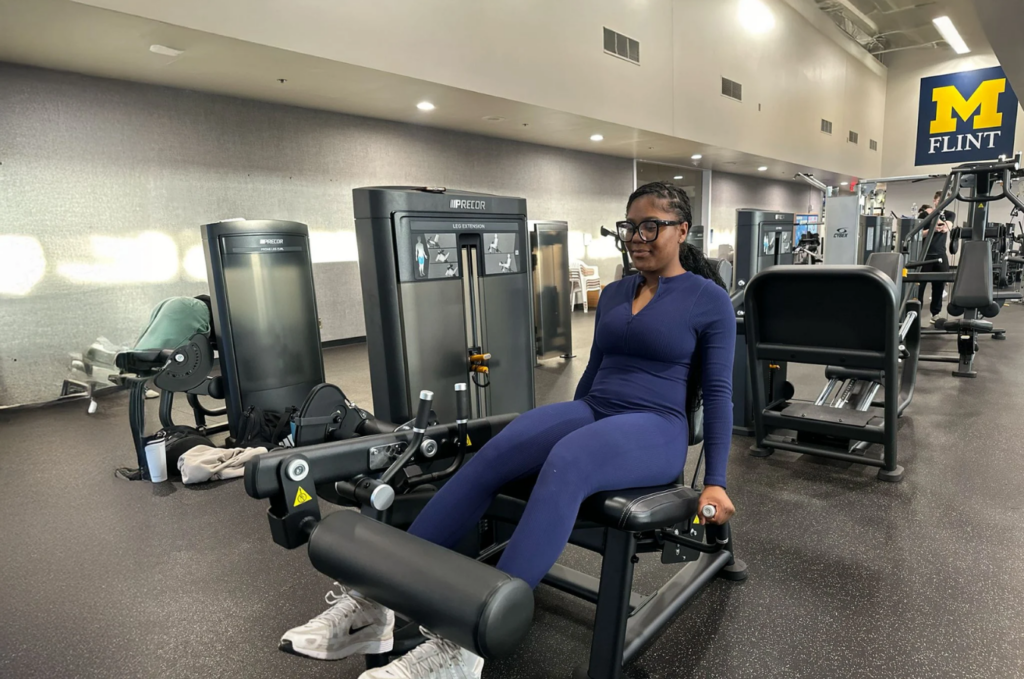 A person is seated on a leg extension machine in a gym, with exercise equipment and a "M FLINT" sign visible in the background.