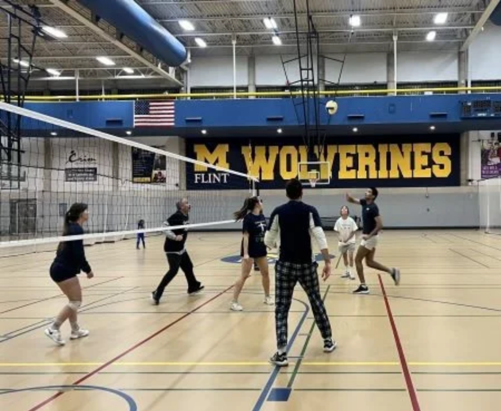 A group of people playing volleyball in a gym with a large "M WOLVERINES" banner on the wall.