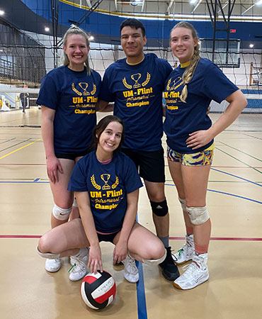 A group of four volleyball players in matching blue shirts pose together on a gym court, holding a volleyball.