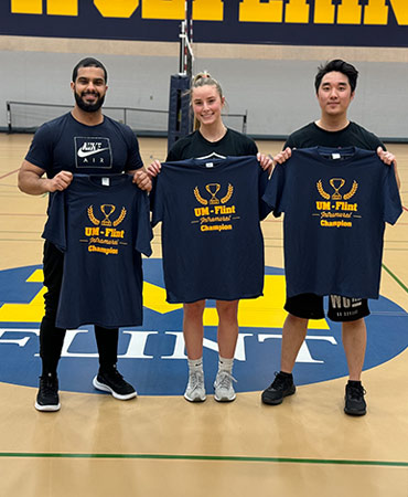 Three people stand on a gym floor holding navy blue t-shirts that read "UM-Flint Champion."