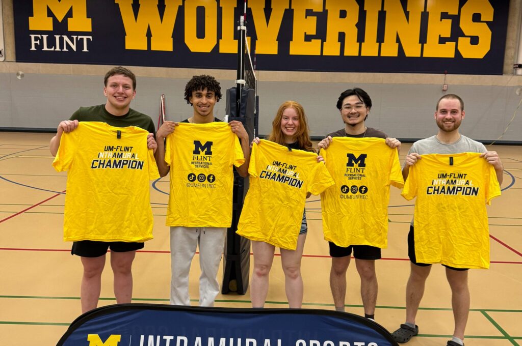 Four people hold yellow t-shirts that say "UM-FLINT INTRAMURAL CHAMPION" in a gym with a "Wolverines" banner in the background.