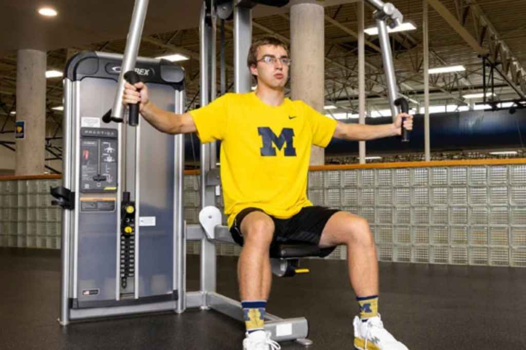 A person using a chest press machine in a gym, wearing a yellow shirt with a large letter "M" and black shorts.