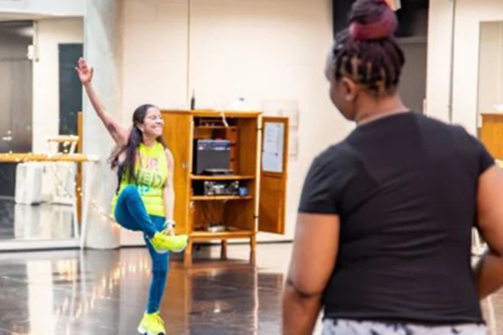 A person in a bright outfit is dancing with one arm raised, while another person watches from behind in a studio setting.