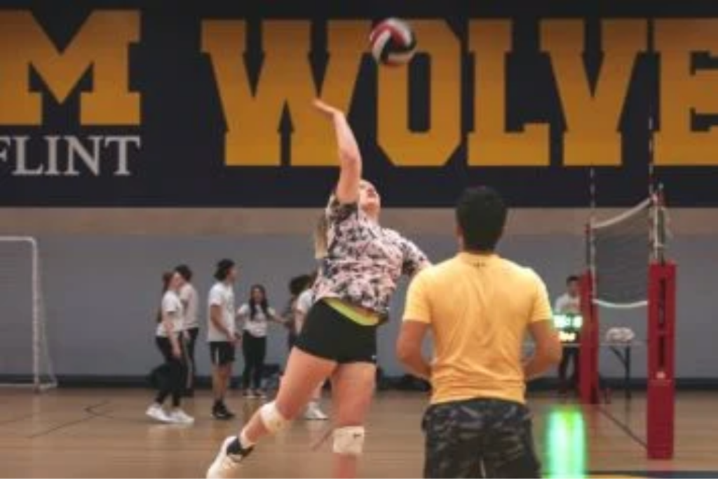 A person serves a volleyball in a gym, with a group of people watching in the background.