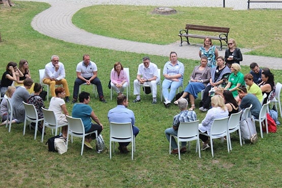 A large group of people seated in a circle of chairs outdoors, engaged in discussion.