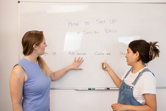 Two people at a whiteboard discussing steps to set up a social wall: sign up, add sources, customize, and share.