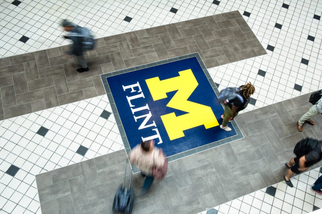 Aerial view of a tiled floor with the University of Michigan Flint logo, as people walk across it.