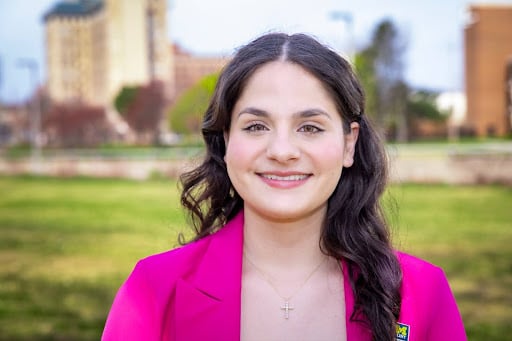 A person smiles in a pink blazer, standing outdoors with a park and buildings in the background.