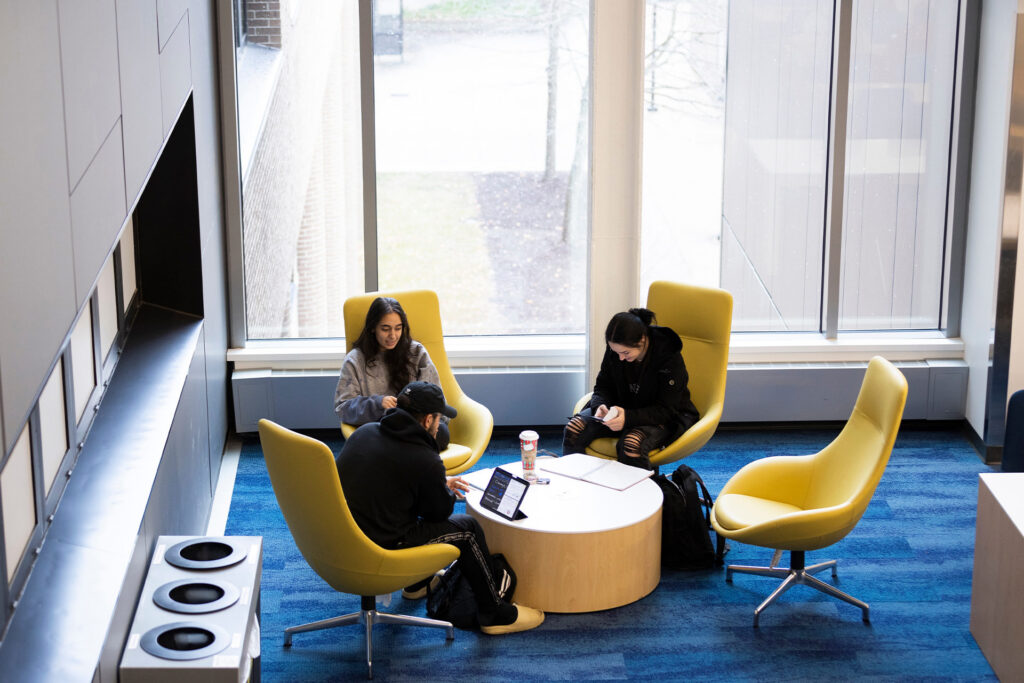 Three people are seated in yellow chairs around a circular table, engaged in conversation and using devices, in a well-lit indoor space.