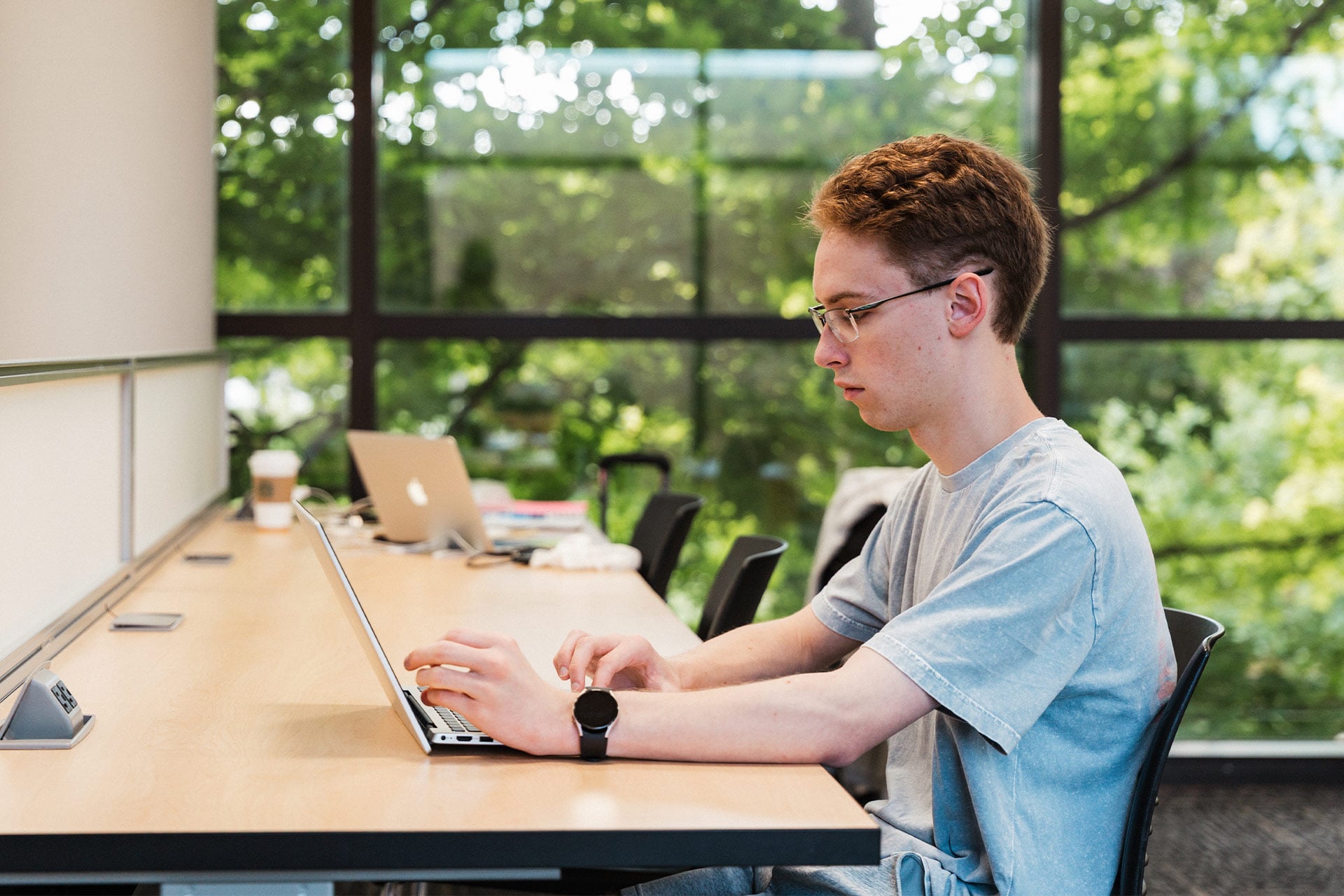 A person sitting at a wooden table, using a laptop, with a coffee cup and other items on the table, surrounded by greenery outside.