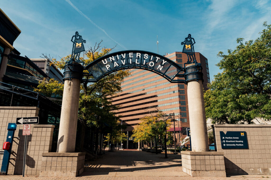 Archway sign reading "UNIVERSITY PAVILION" with a blue sky and buildings in the background, surrounded by trees.
