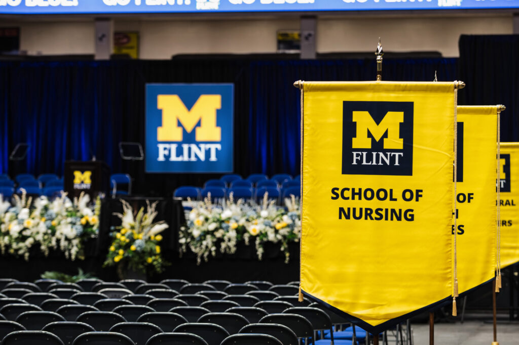 A graduation ceremony setup featuring yellow banners for the School of Nursing and University of Michigan-Flint, with floral arrangements and empty chairs.