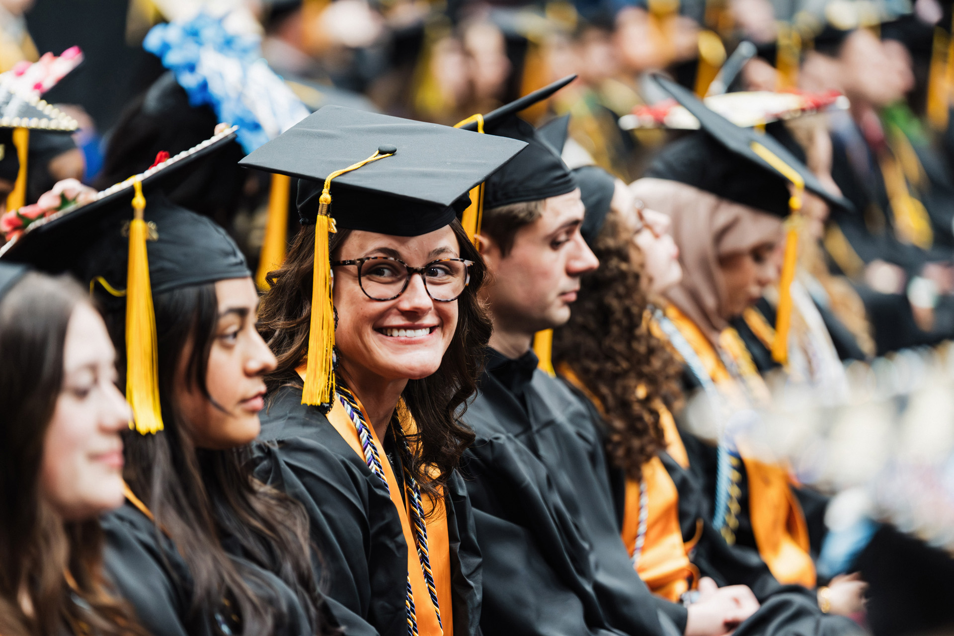 A group of graduates wearing black caps and gowns, some with yellow tassels, seated in a ceremony.