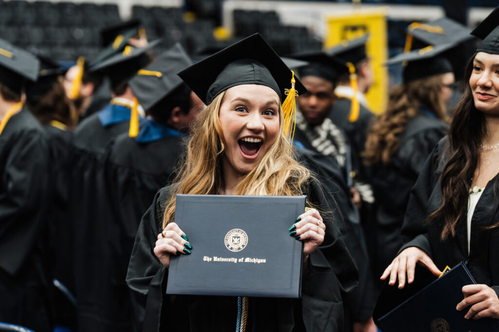 A person in a graduation cap and gown holds a diploma from the University of Michigan, surrounded by other graduates.