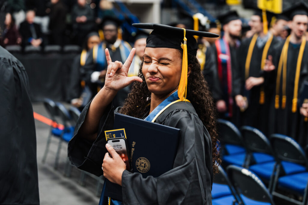 A graduate in a cap and gown holds a diploma and makes a hand gesture while standing among other graduates at a ceremony.