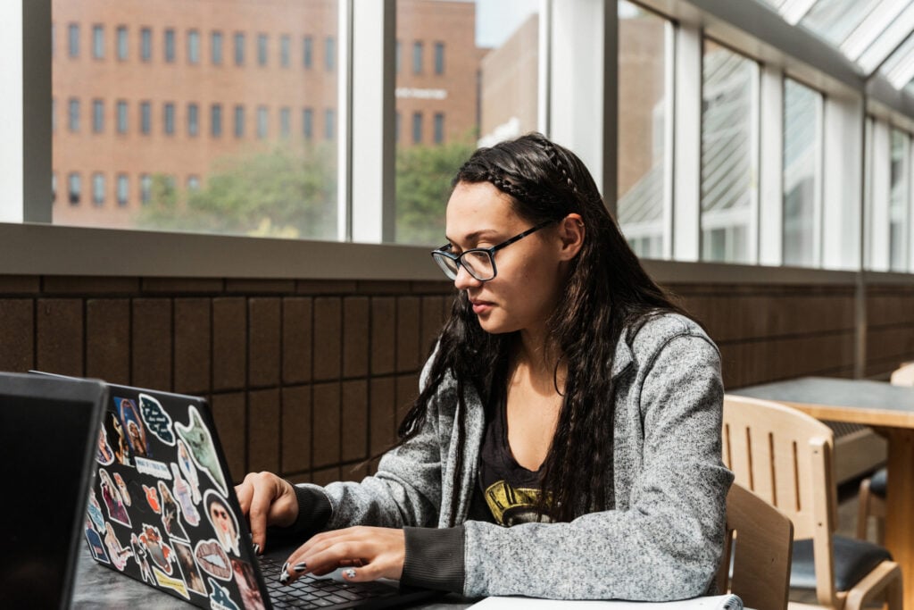 A person sits at a table using a laptop covered in stickers, with large windows and a view of a building in the background.