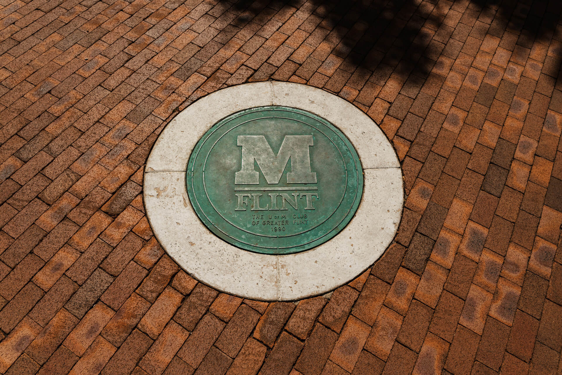A circular green metal plaque embedded in a brick pavement, featuring the text "M FLINT" and "THE JAYCEES OF GREATER FLINT 1980."