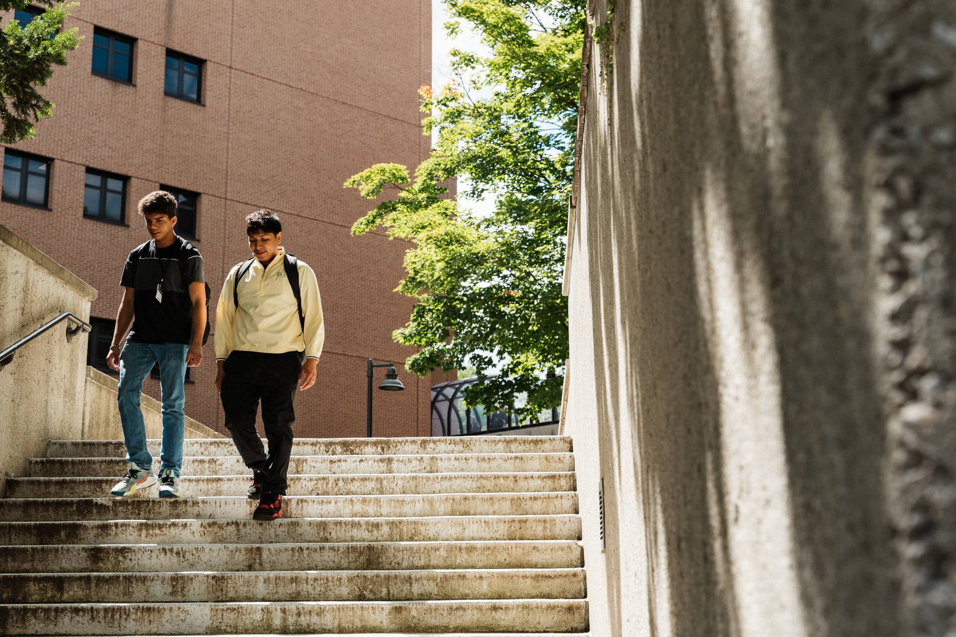 Two individuals walk down a set of outdoor stairs, surrounded by greenery and a brick building in the background.