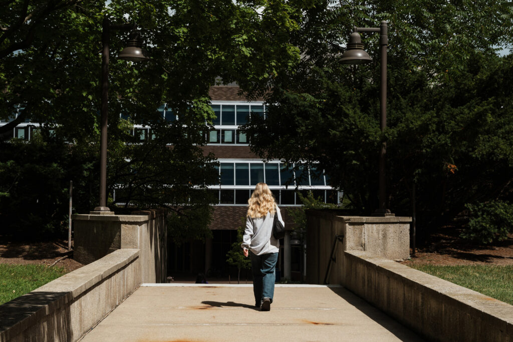 A person with long, wavy hair walks down a concrete path towards a building surrounded by trees and grass.