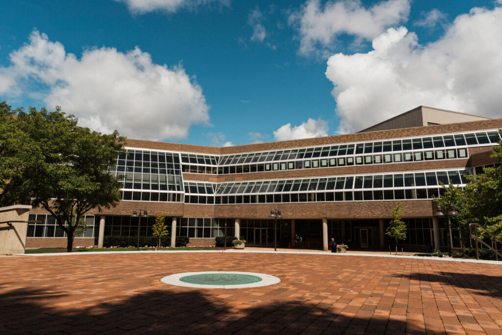 A modern building with large glass windows, surrounded by trees and a brick-paved courtyard under a blue sky with clouds.