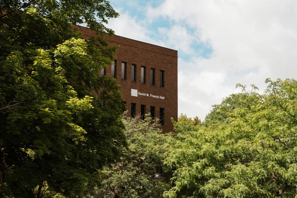 A brick building partially obscured by green trees, featuring a sign that reads "David M. French Hall."