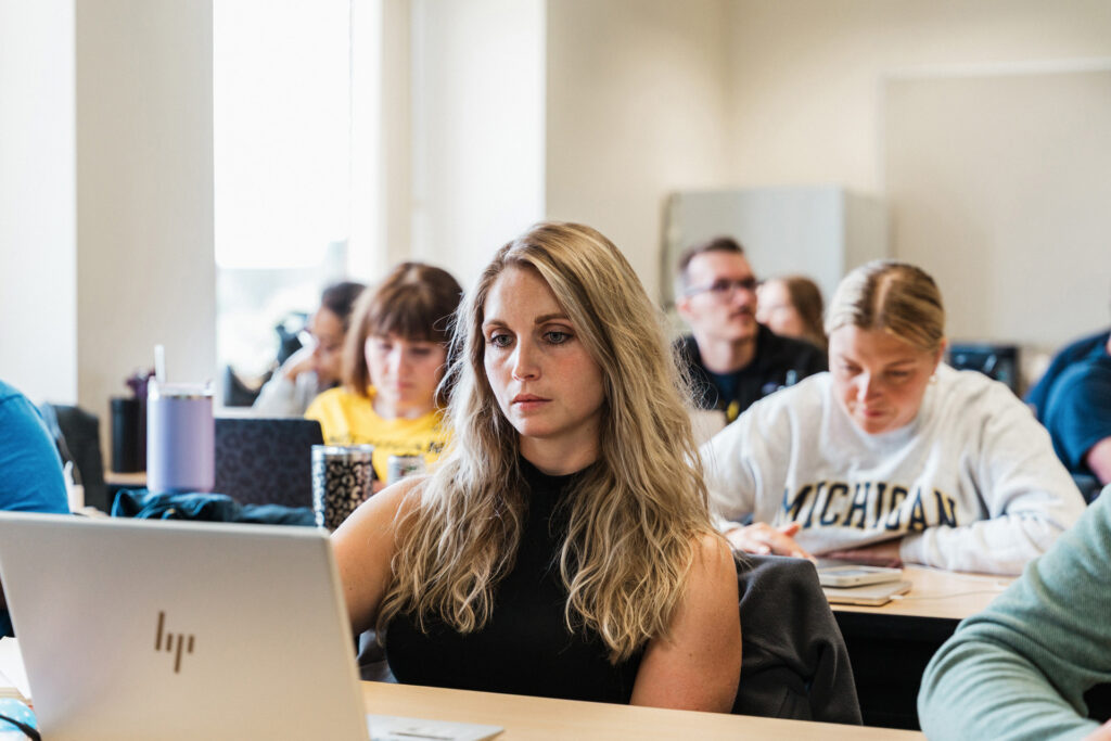A person sits at a desk with a laptop, focused on a classroom setting with several other students in the background.