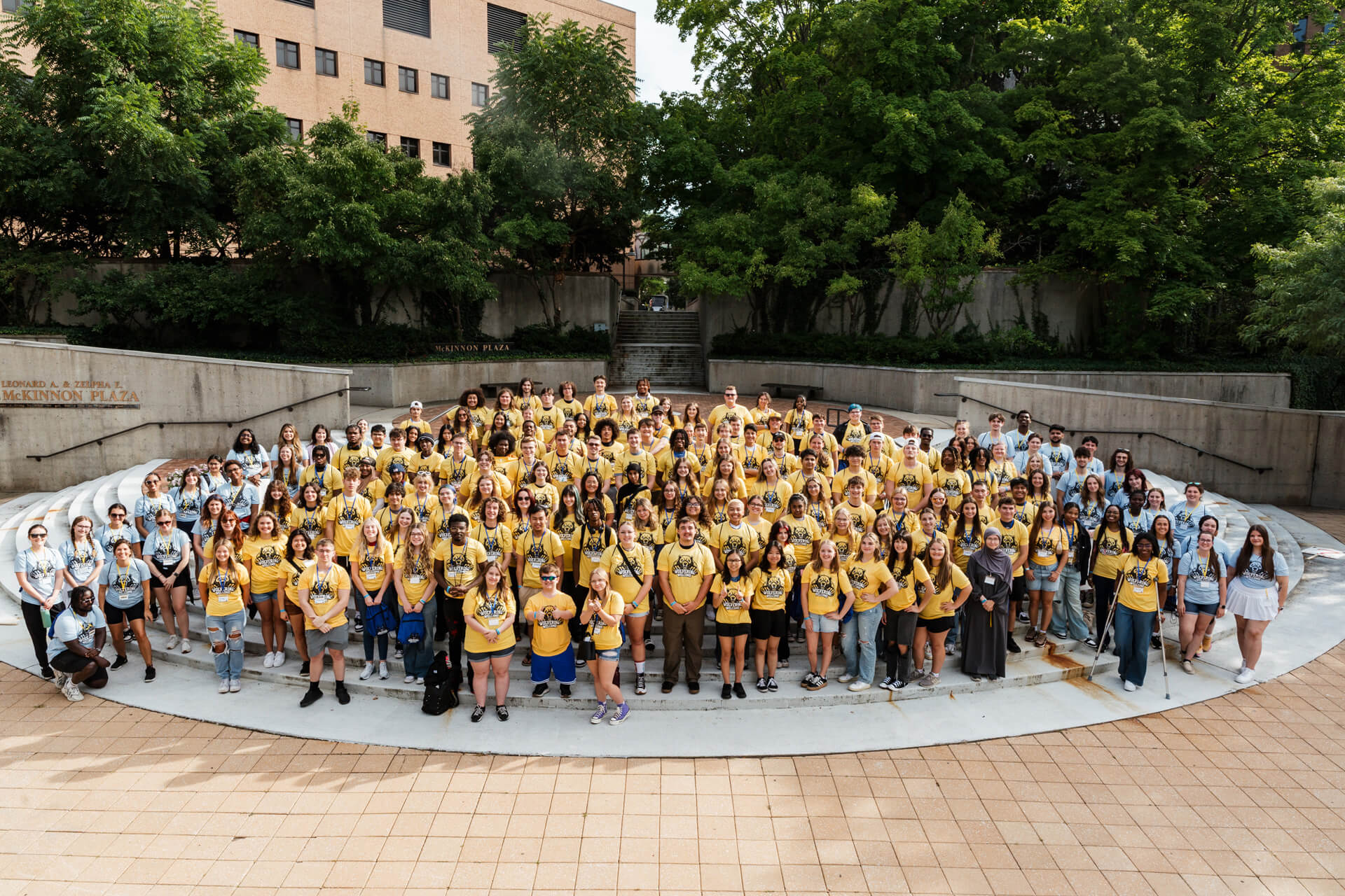 A large group of students wearing yellow t-shirts stands together in McKinnon Plaza, surrounded by greenery and buildings.