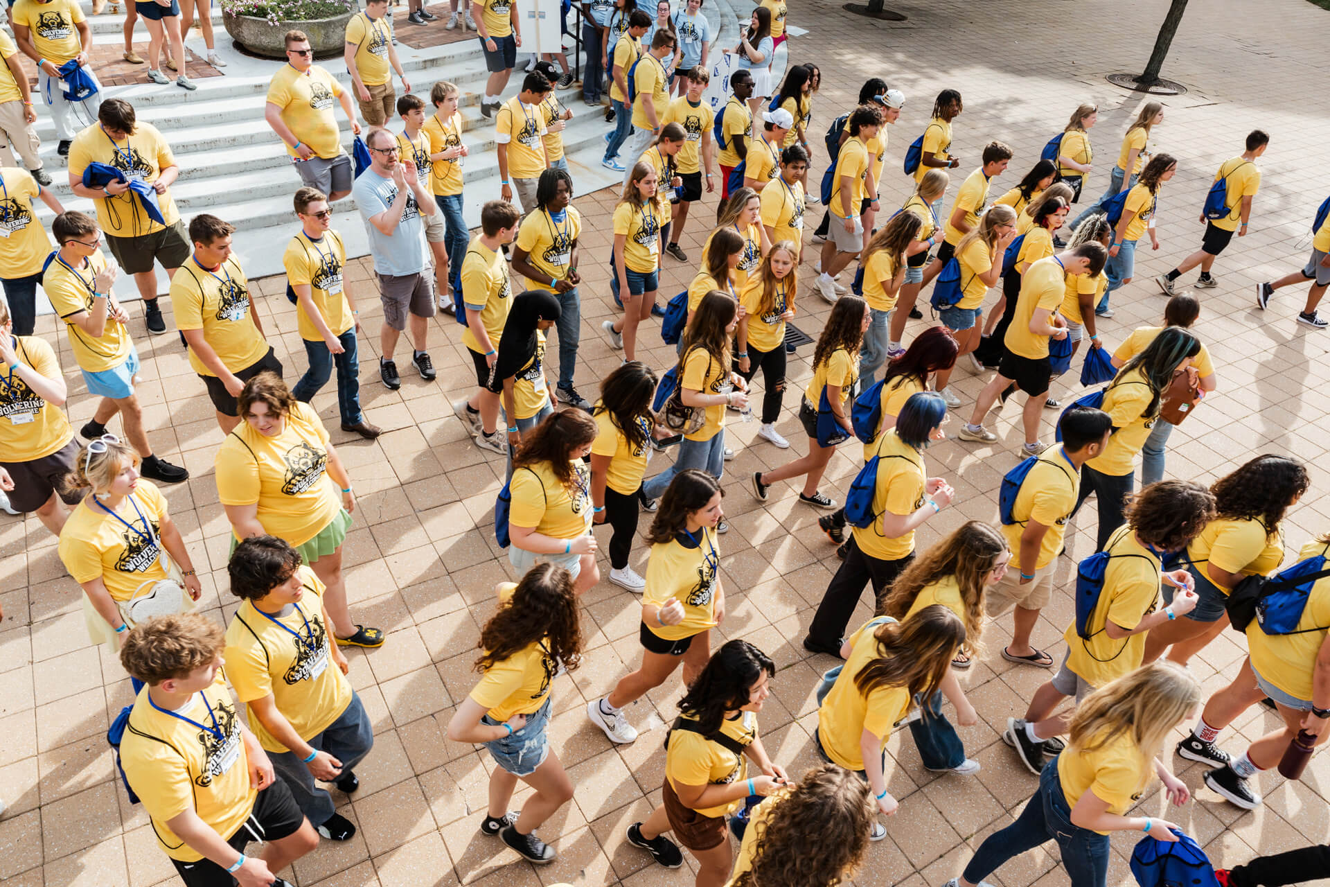 A large group of people wearing yellow t-shirts and carrying blue bags are walking together on a paved area, with some stairs in the background.