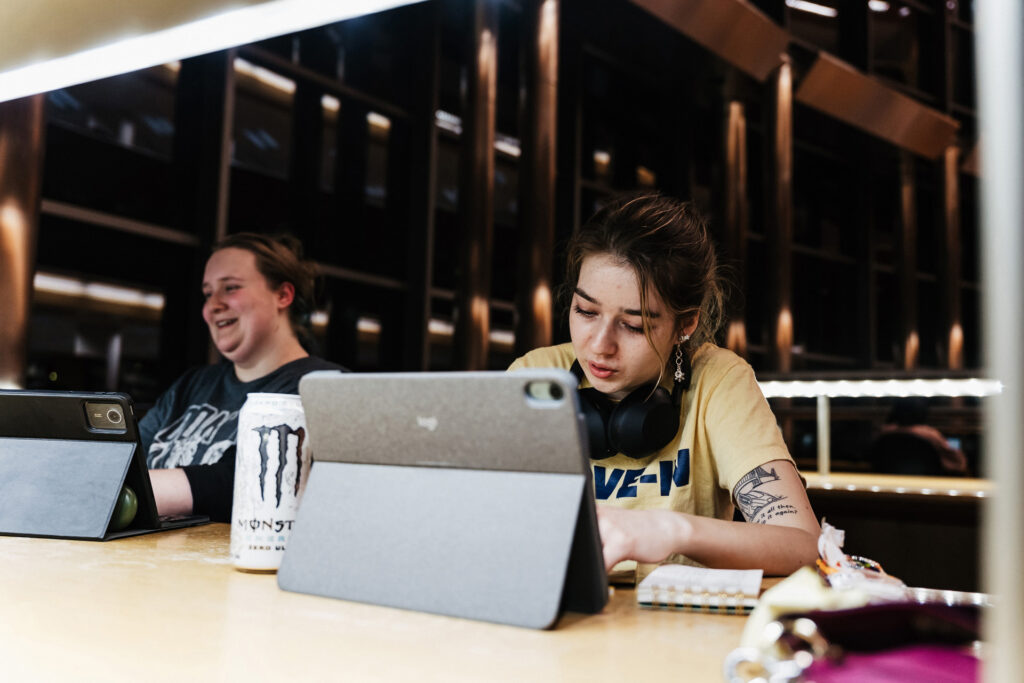 Two people are seated at a table with tablets, one person is writing in a notebook, and a can of Monster energy drink is visible.