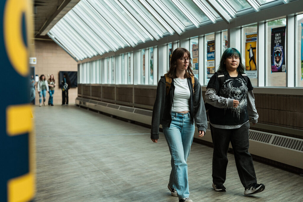 Two students walk down a brightly lit hallway with large windows, passing by posters on the walls.