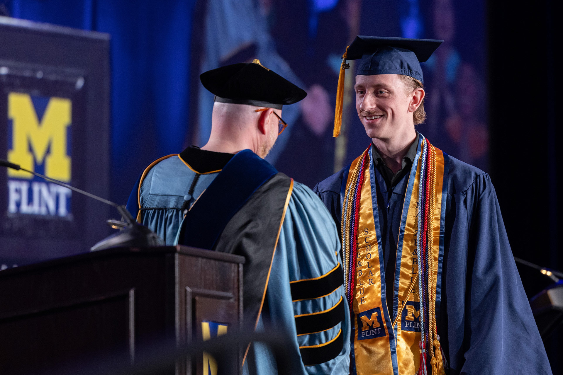 A graduate in a blue cap and gown, wearing multiple honor cords, stands on stage receiving recognition from a faculty member in academic regalia. A banner with "M FLINT" is visible in the background.