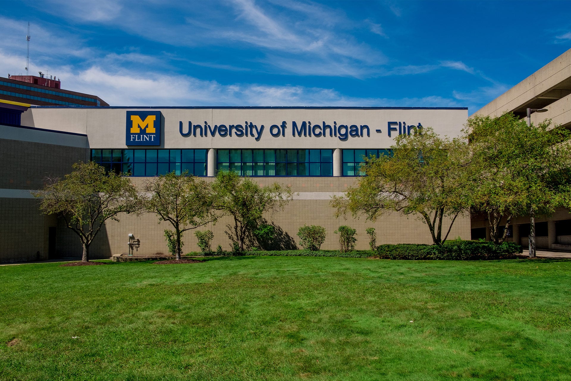 The image shows the exterior of the University of Michigan-Flint building, featuring a blue sky, green grass, and trees in the foreground.