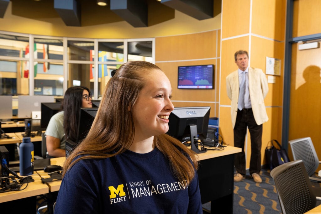 A student wearing a "M FLINT SCHOOL OF MANAGEMENT" shirt sits in a computer lab, with a professor standing in the background.
