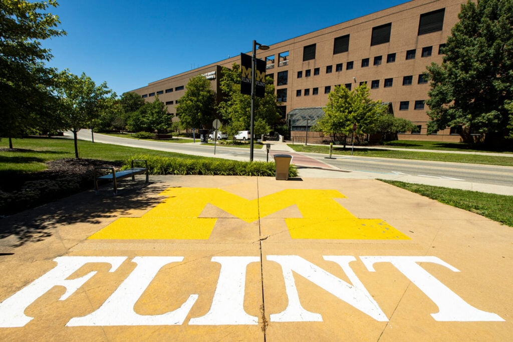 A concrete pathway with a large yellow "M" and the word "FLINT" in white, surrounded by trees and a building in the background.