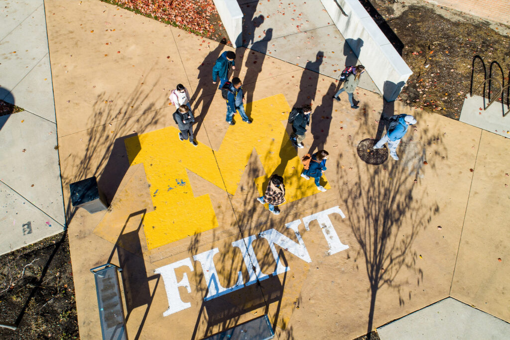 Aerial view of a group of people walking on a pathway with a large yellow "M" and the word "FLINT" painted on the ground.